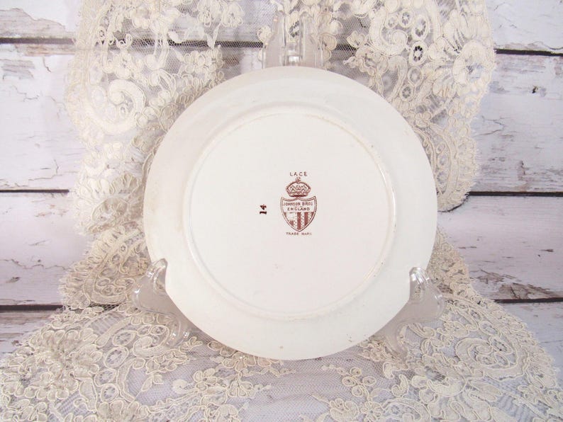Pu&ograve; includere: Un piatto in ceramica bianca con un emblema marrone e il testo "LACE" e "JOHNSON BROS ENGLAND". Il piatto &egrave; su un supporto trasparente, con una tovaglia di pizzo sullo sfondo.