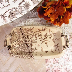 May include: A rectangular, off-white ceramic serving tray with brown floral and bamboo designs. The tray has two handles and is decorated with a variety of patterns. A second tray is partially visible in the background.
