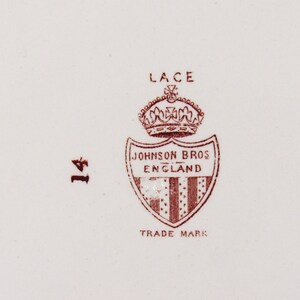 Pu&ograve; includere: Primo piano di un piatto in ceramica bianca con un emblema marrone. L'emblema presenta una corona sopra uno scudo con la scritta "JOHNSON BROS ENGLAND" e "TRADE MARK". La parola "LACE" &egrave; sopra la corona e il numero "14" &egrave; a sinistra.