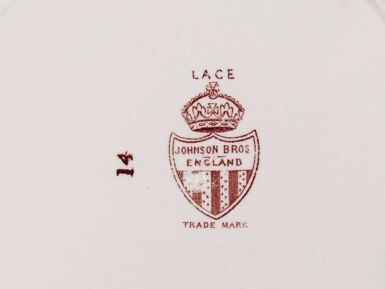 Pu&ograve; includere: Primo piano di un piatto in ceramica bianca con un emblema marrone. L'emblema presenta una corona sopra uno scudo con la scritta "JOHNSON BROS ENGLAND" e "TRADE MARK". La parola "LACE" &egrave; sopra la corona e il numero "14" &egrave; a sinistra.