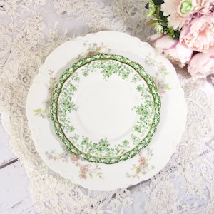 May include: A white ceramic saucer featuring a green floral pattern along the scalloped edge, layered on top of a larger white plate with a faint pink floral design. The saucer has a gold-trimmed rim and a central white circular area.