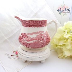May include: A white and red ceramic pitcher with a landscape design. The pitcher is placed on a small, white, round stand with three feet. The pitcher has a handle and a spout.