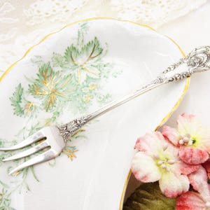 May include: A silver fork with an ornate handle rests on a white dish with a gold rim and green floral design. Pink and white flowers are in the foreground. The fork has three tines and detailed engravings.