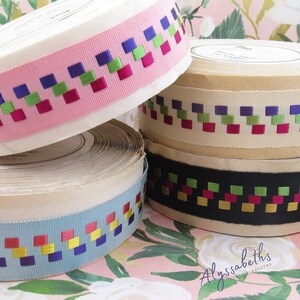 May include: Four spools of ribbon with a geometric pattern of squares in pink, cream, blue, and black. The ribbon has a woven design with squares in shades of green, purple, red, and yellow.