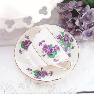 May include: A white teacup and saucer with a purple violet floral pattern. The saucer has a gold trim.