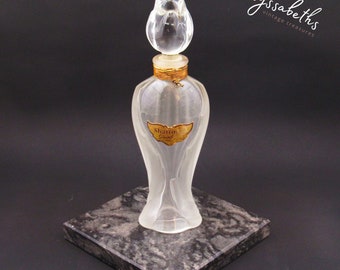 May include: A clear glass perfume bottle with a rose-shaped stopper. The bottle has a gold label that reads "Shazim Grand". The bottle is sitting on a black marble base.