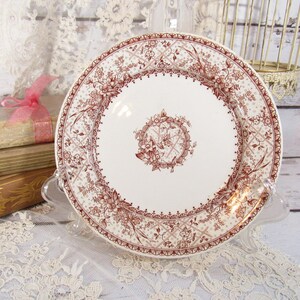 Pu&ograve; includere: Un piatto in ceramica bianca con un dettagliato motivo floreale e geometrico marrone. Il piatto &egrave; esposto su un supporto trasparente, con libri e pizzo sullo sfondo. Il piatto ha un diametro di circa 25 cm.