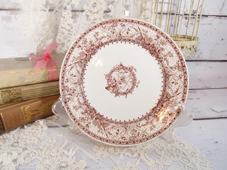 Pu&ograve; includere: Un piatto in ceramica bianca con un dettagliato motivo floreale e geometrico marrone. Il piatto &egrave; esposto su un supporto trasparente, con libri e pizzo sullo sfondo. Il piatto ha un diametro di circa 25 cm.