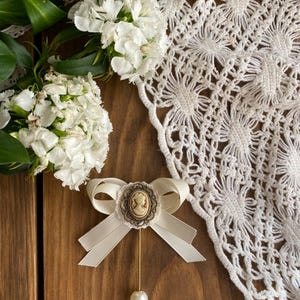May include: A white satin bow brooch with a cameo featuring a woman's profile. The brooch is accented with a pearl and a gold pin.