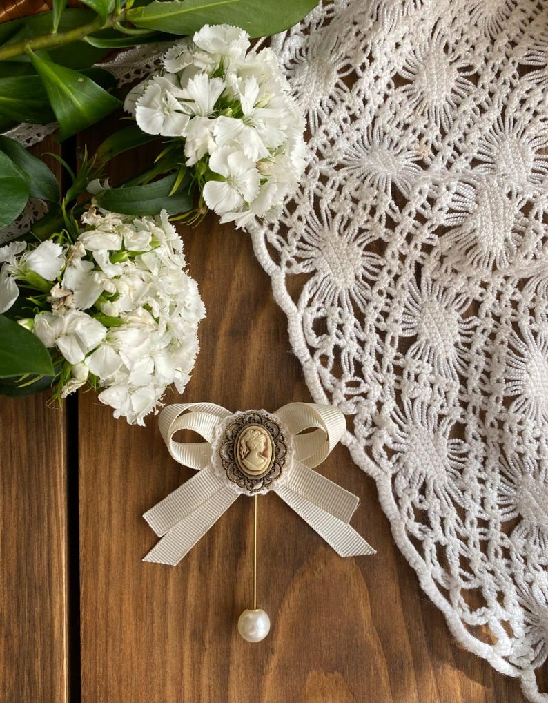 May include: A white satin bow brooch with a cameo featuring a woman's profile. The brooch is accented with a pearl and a gold pin.