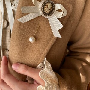 May include: A white satin bow with a cameo brooch and a pearl pin on a brown fabric background.