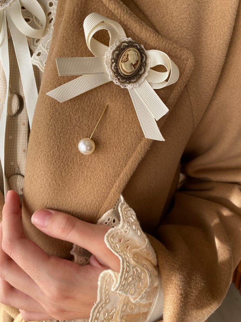 May include: A white satin bow with a cameo brooch and a pearl pin on a brown fabric background.