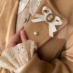 May include: A white satin bow brooch with a cameo and pearl pin. The brooch is pinned to a brown coat.