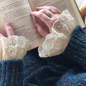 May include: A close-up of a person's hands holding a book with a blue and white cover. The person is wearing a blue sweater with white lace trim on the cuffs. The text on the book reads "Ayet Bir Akşam..."