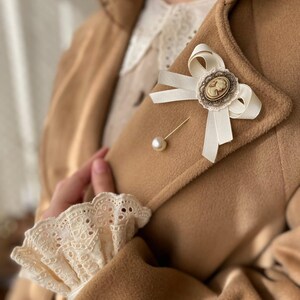 May include: A white satin bow with a cameo brooch pinned to a brown coat. The bow is tied with a pearl pin.