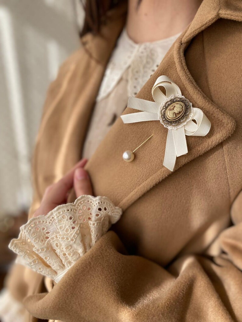 May include: A white satin bow with a cameo brooch pinned to a brown coat. The bow is tied with a pearl pin.