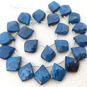 Puede incluir: Un hilo de cuentas de azurita azul, facetadas y de forma irregular tipo escudo, con inclusiones de matriz marrón oscuro. Las cuentas están ensartadas en un hilo azul claro y separadas por pequeñas cuentas espaciadoras de metal plateado.