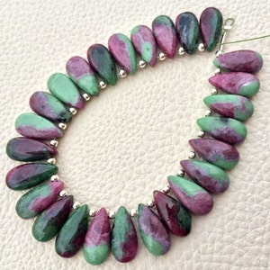 Może przedstawiać: Sznur koralików w kształcie łezki. Każdy koralik to mieszanka kolorów zielonego i bordowego, o wypolerowanej powierzchni. Koraliki są nawleczone małymi srebrnymi koralikami pomiędzy każdą łezką. Koraliki leżą na białej powierzchni.