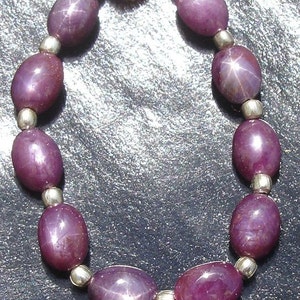 Könnte beinhalten: Eine Halskette mit ovalen, tiefvioletten Edelsteinen mit Sterneneffekt, aufgereiht mit kleinen silbernen Perlen. Der Verschluss ist aus goldfarbenem Metall. Die Edelsteine sind etwa 1,3 cm lang.