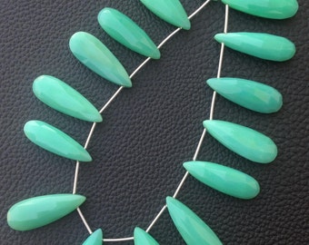 May include: A strand of teardrop-shaped, faceted, light green gemstones. The beads are strung on a thin, silver-colored wire, arranged in a curved pattern. The background is a dark, textured surface.