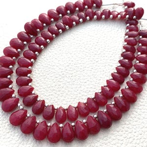 May include: A necklace featuring two strands of faceted, teardrop-shaped, deep red gemstones. The beads are strung with small silver-colored spacers. The necklace is displayed on a white surface.