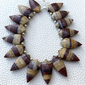 Puede incluir: Un collar de cuentas de piedras preciosas. Cada cuenta tiene forma de punta de flecha alargada y facetada, con bandas alternas de color beige, crema y burdeos. Las cuentas están ensartadas en un alambre plateado.