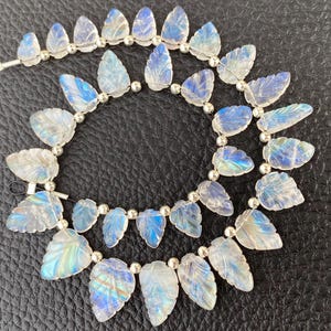 May include: A strand of translucent, leaf-shaped moonstone beads with a blue iridescent sheen, interspersed with small, round, polished silver-coloured metal spacer beads. The carved gemstone leaves are arranged in a circular pattern on a string.