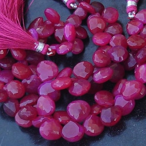 Pode incluir: Um fio de contas facetadas em forma de coração, em tons de magenta e vermelho rubi. As contas são enfiadas juntas, com uma borla de fio magenta a condizer no topo. As contas têm aproximadamente 1 cm.