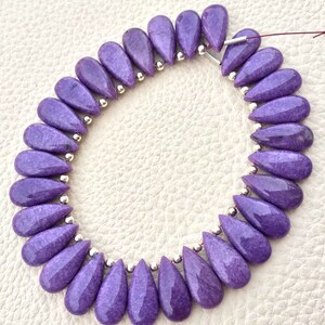 Puede incluir: Un collar de cuentas púrpuras pulidas, facetadas, en forma de lágrima. Cada cuenta está conectada por pequeñas cuentas de color plateado. Las cuentas están ensartadas en un hilo fino de color marrón rojizo, creando un collar o pulsera.