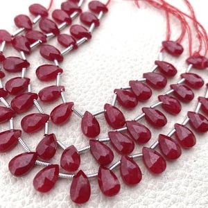 Puede incluir: Tres hilos de cuentas de piedras preciosas de color rojo intenso, talladas en facetas y con forma de pera, dispuestas en forma curva. Cada cuenta está separada por pequeños espaciadores metálicos plateados y los hilos se unen con hilos rojos finos.