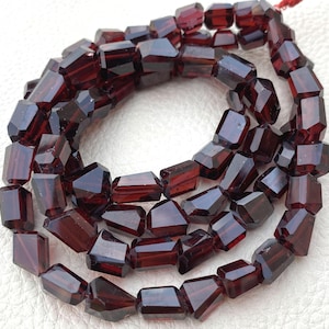 May include: Strand of faceted, dark red gemstone beads. The beads have a rectangular shape with multiple facets, creating a sparkling effect. The beads are strung together on a red string, ready for jewelry making.