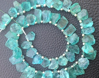 Puede incluir: Un collar de piedras preciosas de color azul verdoso claro, cada una con bordes irregulares y una calidad translúcida. Las piedras están ensartadas con pequeñas cuentas plateadas. La disposición se muestra sobre un fondo oscuro y texturizado.