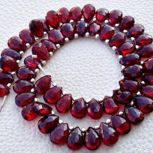 May include: Strands of faceted, teardrop-shaped, deep red garnet beads. The beads are strung together with small silver-colored spacers. The gemstones have a polished, reflective surface, and are arranged in a curved pattern.