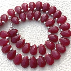 May include: Strands of faceted, teardrop-shaped, deep red gemstones strung together with silver-colored beads. The gemstones have a polished, reflective surface, and are arranged in a curved pattern.