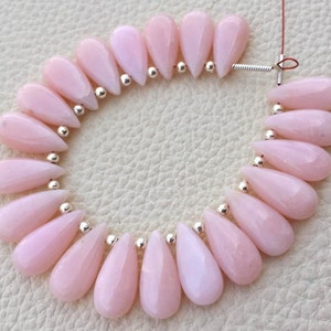 May include: A strand of pale pink teardrop-shaped beads with faceted surfaces. Small silver-colored beads separate each pink bead. The beads are strung on a thin wire, ready for jewelry making. The beads are a soft, pastel color.