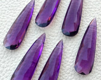 May include: Six faceted, teardrop-shaped amethyst gemstones. Each gem is a deep purple color with a pointed top and a rounded base. The gems are arranged on a white surface.