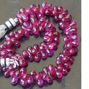 Puede incluir: Un collar de cuentas de piedras preciosas facetadas, de color magenta intenso. Las cuentas tienen forma de lágrima y están ensartadas, con una borla plateada en el extremo. Las cuentas están dispuestas en un patrón en espiral.