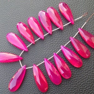 May include: A strand of faceted, teardrop-shaped beads in a vibrant magenta hue. The beads are strung together on a thin, white string, showcasing their polished surfaces and geometric cuts. The beads are approximately 1.5 inches long.