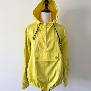 Könnte beinhalten: Eine leuchtend gelbe Kapuzenjacke mit glänzendem, wasserabweisendem Finish. Sie verfügt über einen halben Reißverschluss mit rundem Zipper, eine große Fronttasche mit zwei silberfarbenen Druckknöpfen und schwarze Kordelzüge an Kapuze und Saum.