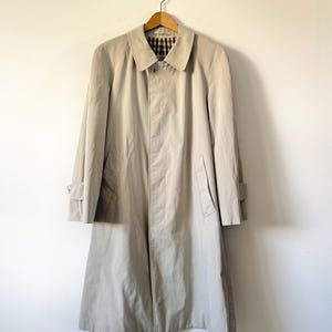 May include: A beige trench coat suspended from a wooden hanger. The coat features a traditional design with a collar, pockets, and button fastenings. The collar's interior lining displays a brown and tan plaid pattern. The coat is crafted from a lightweight fabric.