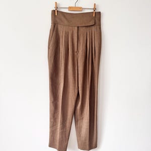 Puede incluir: Pantalones marrones de cintura alta con una cinturilla ancha y pliegues. Los pantalones cuelgan de una percha de madera con pinzas metálicas sobre un fondo blanco. Los pantalones tienen un corte a medida y un estilo clásico.