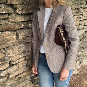 Pode incluir: Um blazer xadrez marrom e branco com botões dourados é usado sobre uma camiseta branca e jeans azuis. Uma bolsa de couro marrom com uma alça de corrente dourada é segurada. A roupa é casual e elegante.