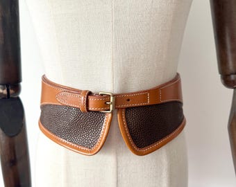 May include: A brown leather belt with a gold-colored buckle. The belt has a textured, dark brown center section and a lighter brown border with white stitching. The belt is displayed on a mannequin.