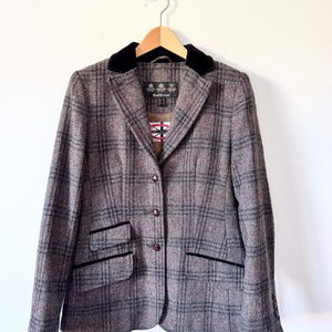 Puede incluir: Blazer de tweed marrón y gris con cuello y ribetes de bolsillo de terciopelo negro. La chaqueta, con un patrón de cuadros, está abotonada y colgada de una percha de madera. La etiqueta de la marca es visible.