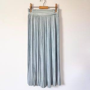 May include: A light blue pleated skirt hanging on a wooden hanger. The skirt has a wide waistband and falls to the ankles. The fabric has a soft, flowing appearance, and the pleats run vertically down the length of the skirt.