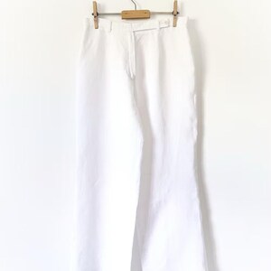 May include: White linen pants hanging on a wooden hanger with clips. The pants have a straight leg and a relaxed fit.