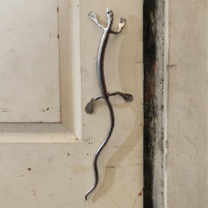 May include: A silver metal lizard door handle with a long tail and four legs. The handle is mounted on a white wooden door.