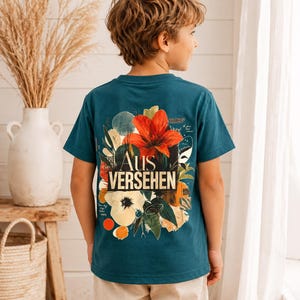 Könnte beinhalten: Ein blaugrünes Kurzarm-T-Shirt mit einer großen, farbenfrohen Blumen-Grafik auf der Rückseite. Das Design zeigt eine markante orangefarbene Lilie, eine weiße Blume und abstrakte Formen mit dem Text "Aus VERSEHEN" in fetter Schrift in der Mitte.