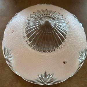 May include: A vintage frosted glass light fixture shade. The shade has a textured surface and a clear glass floral design in the center. The edges feature decorative floral accents.