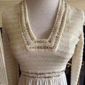 Puede incluir: Vestido vintage color marfil con mangas largas. El corpiño presenta un escote en V y una cintura adornada con pequeños detalles redondos dorados. La falda es de tela blanca lisa y fluida.